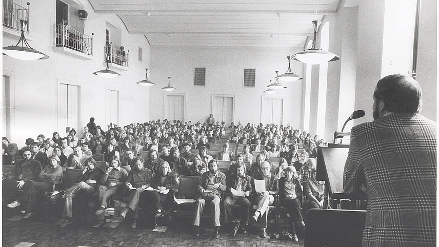 A black and white shot of a lecture in a large hall. In the foreground, a speaker stands at a lectern while many members of the audience sit in rows and look towards him. Some are holding documents in their hands.