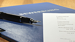 A blue application folder contains a black pen and a printed CV.