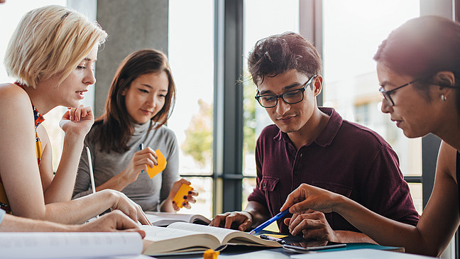 Five students of different nationalities sit together at a table in front of open books and confer with sticky notes in their hands.
