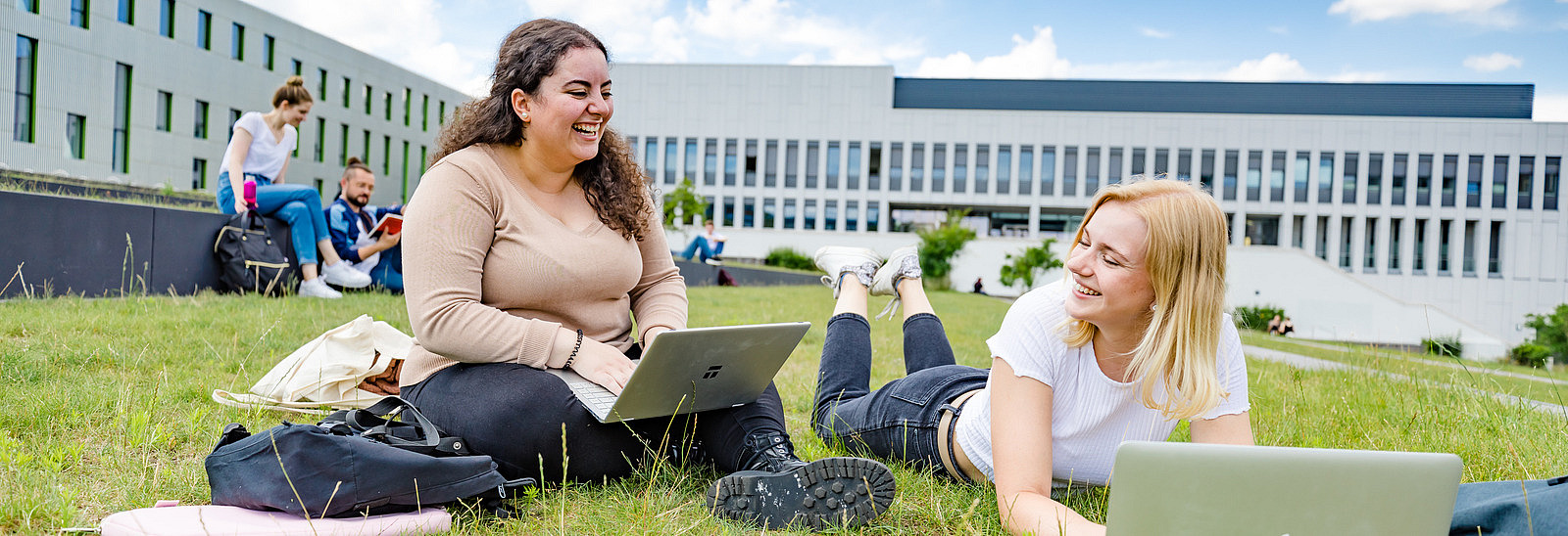 Zwei Studentinnen auf dem begrünten Platz neben der Bibliothek. Eine von ihnen sitzt, die andere liegt auf dem Rasen, beide haben Laptops vor sich