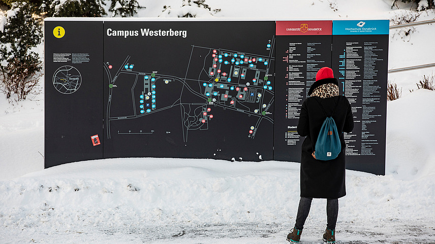 A person in winter clothing stands in front of a large wall with a map of the campus on Westerberg. There is snow.