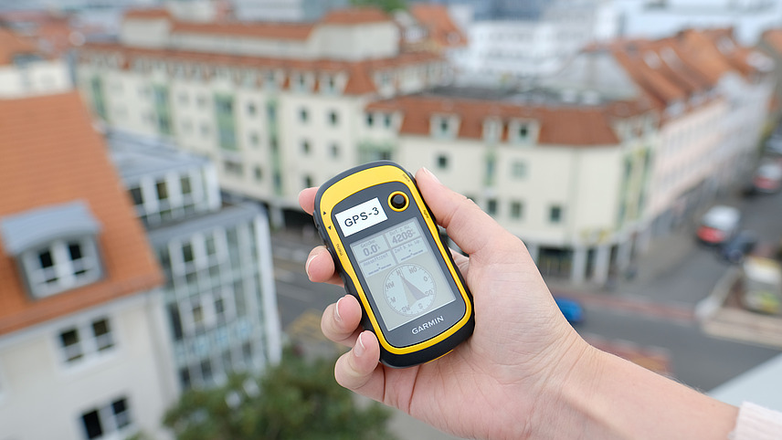 A GPS device is held over the edge of a railing. A bird's eye view of an intersection in Osnabrück can be seen in the background. 