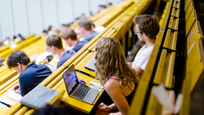 Side view of yellow rows of seats in a lecture hall. A female student sits leaning forward in front of her open laptop with her gaze directed forward while the person sitting next to her looks at his notes. Other students are sitting around them in the rows of seats.